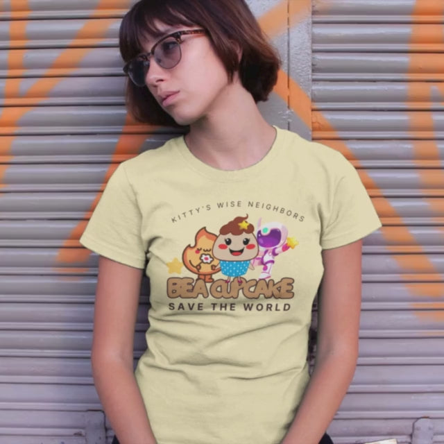 Woman in yellow t-shirt, leaning against a gray garage door with orange graffiti. The t-shirt reads "be a cupcake, save the world" with hello kitty like cartoons but 3 cupcakes. Symbolizing the 3 life modes: exist, flow and transcend