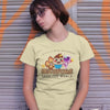 Woman in yellow t-shirt, leaning against a gray garage door with orange graffiti. The t-shirt reads "be a cupcake, save the world" with hello kitty like cartoons but 3 cupcakes. Symbolizing the 3 life modes: exist, flow and transcend